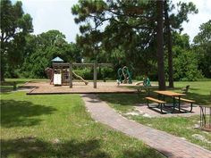 an empty park with picnic tables and play equipment in the distance, surrounded by trees