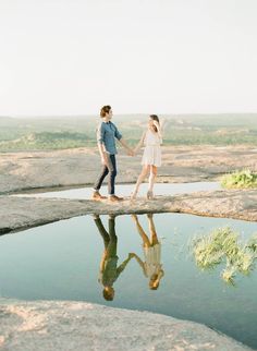 two people holding hands and walking across a bridge over a body of water in the desert