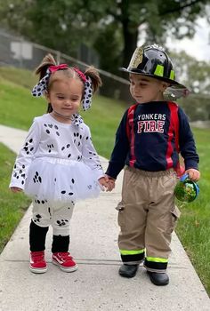 two young children dressed in fireman's gear walking down a sidewalk holding hands