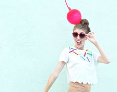 a young woman wearing sunglasses and holding a red ball above her head, standing in front of a blue wall