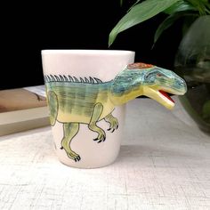 a mug with a dinosaur painted on it sitting next to a potted plant and books