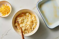 two bowls filled with shredded cheese next to a baking pan and wooden spoon on a marble counter top