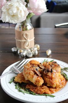 a white plate topped with meat covered in sauce next to a vase filled with flowers