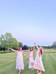 three women in dresses are holding hands and dancing on the green grass with trees in the background