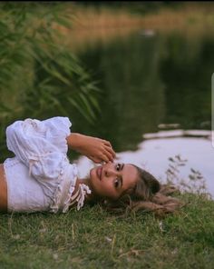 a woman laying on the grass next to a body of water wearing a white dress