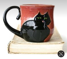 a black cat sitting on top of a red coffee cup next to an open book