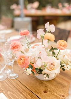 an arrangement of flowers in a white bowl on a wooden table with wine glasses and napkins