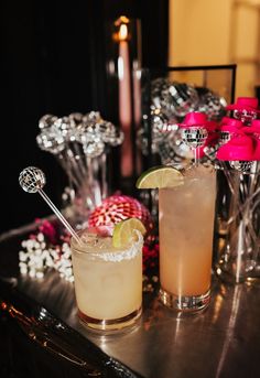 two cocktails are sitting on a table with pink and silver decorations in the background