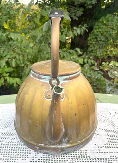 an old fashioned tea kettle sitting on top of a white tablecloth with trees in the background