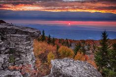 the sun sets over mountains and trees on top of a rocky cliff in the fall