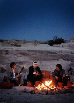 three people sitting around a campfire in the desert at night with one person playing guitar