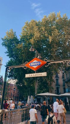 people are walking under the metro sign in front of some buildings on a sunny day