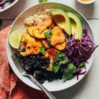 a white plate topped with shrimp, rice and veggies next to an avocado