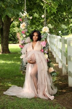 a pregnant woman in a dress sitting on a swing with flowers and greenery around her