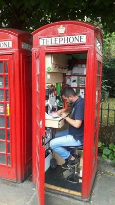 two red telephone booths sitting next to each other on a sidewalk with one man working in the phone booth