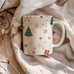 a white coffee mug with christmas decorations on it sitting on a bed next to dried flowers