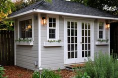 a small gray shed with white windows and plants in the window boxes on each side