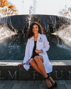 a woman sitting in front of a fountain wearing a white coat and black dress with heels