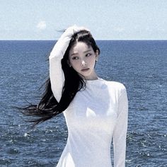 a woman with long black hair standing in front of the ocean wearing a white dress