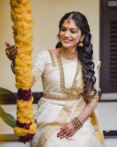 a woman in a white sari and gold jewelry holding a yellow pole with flowers on it