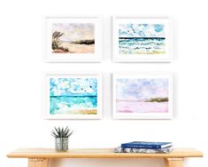 four watercolor paintings hang on the wall above a wooden table with a plant and books