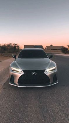 the front end of a silver sports car parked on a road at sunset with its lights on