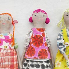 three cloth dolls are standing next to each other on a white wall, one is wearing a colorful dress and the other has pink hair