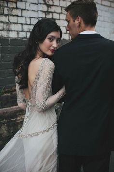 a man and woman standing next to each other in front of a brick wall wearing wedding gowns