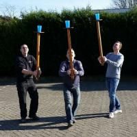 three men holding up wooden poles in the middle of a brick road with grass and bushes behind them