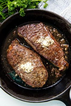 two steaks cooking in a skillet with parsley and garlic on the side