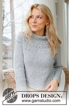 a woman sitting in front of a window wearing a gray knitted sweater and white pants