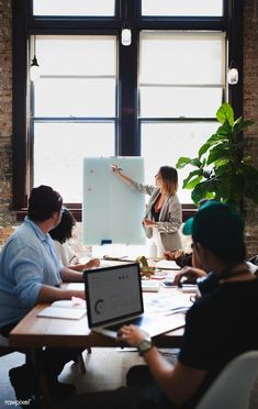 a group of people sitting around a table in front of a whiteboard with a woman pointing at it