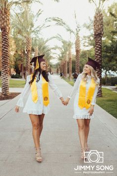 two women in graduation gowns holding hands and walking down the street with palm trees behind them