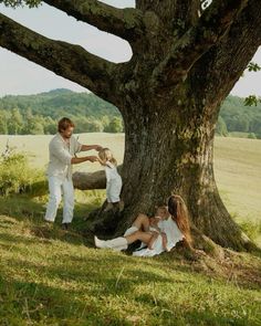 two people sitting on the ground next to a large tree and one person standing in front of it