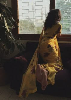 a woman in a yellow kimono sitting on a couch by a window with a potted plant