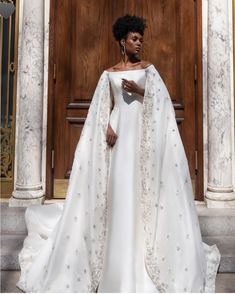 a woman standing in front of a wooden door wearing a white gown and cape with beading on it