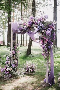 an outdoor wedding ceremony with purple flowers and greenery on the arbor, surrounded by tall trees