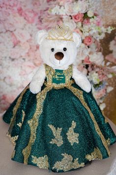 a white teddy bear wearing a green dress and gold tiara with flowers in the background