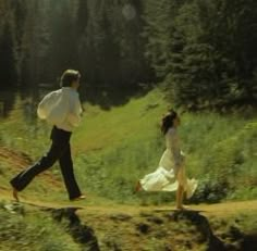 two people are walking on a path in the woods, one is wearing a white dress