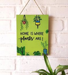 a green sign that says home is where plants are hanging on a brick wall next to a potted plant