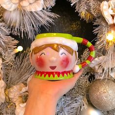 a hand holding a mug with a smiling face on it in front of a christmas tree