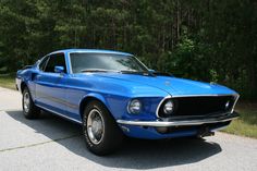 a blue mustang sitting on top of a parking lot next to some trees and grass