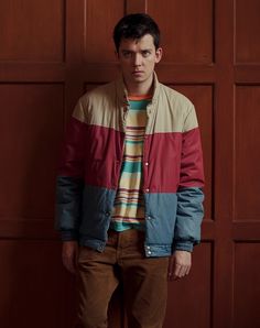 a man standing in front of a wooden wall wearing a colorful jacket and brown pants