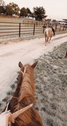 two people riding horses down a dirt road next to a fenced in area with grass