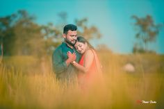 a man and woman standing in tall grass with their arms around each other, looking at the camera