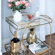 a gold bar cart with wine glasses and bottles on the top, next to a bouquet of pink roses