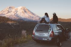 two people sitting on the roof of a car in front of a snow capped mountain