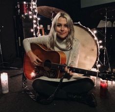 a woman sitting on the floor with a guitar in her lap and lights around her