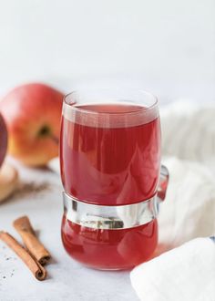 a glass of apple cider next to cinnamon sticks and apples on a white surface