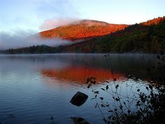 a lake surrounded by trees with fall colors on the mountains in the background and fog hanging over the water